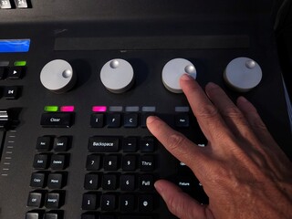 A person operating a professional lighting console panel with one hand, adjusting control knobs and buttons in a studio or live event setting.