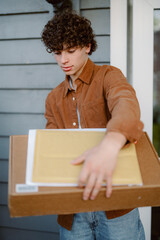 A young individual stands at the entrance of a home, holding a package. They have curly hair and are dressed in a brown shirt. The setting suggests a casual afternoon delivery.