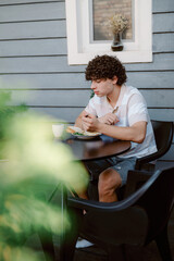 A young man sits at a round table, eating breakfast with a focused expression. Bright green foliage surrounds him, creating a relaxed outdoor atmosphere on a sunny day.