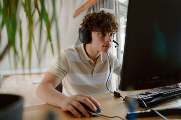 A young boy sits at a wooden desk, focused on his computer screen. He wears a striped polo shirt and a gaming headset. Sunlight streams through a window, adding warmth. © m-art