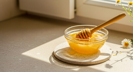 A small glass bowl filled with golden honey sits on a coaster, with a wooden honey dipper resting inside, bathed in warm sunlight near a window with a few daisies.