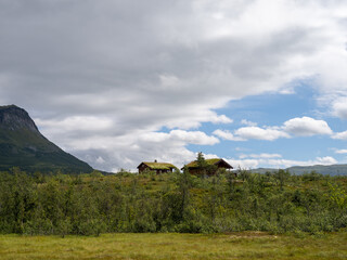 Fototapeta premium Traditional wooden cabins with grass roofs in remote mountain landscape under dramatic clouds and blue sky, concept of eco-friendly living and rural tourism.