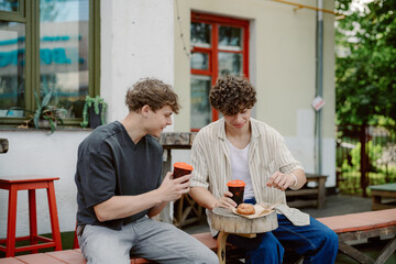Two young men sit at a wooden table in an outdoor cafe, sharing laughs while sipping coffee and enjoying pastries. Greenery and a cozy atmosphere enhance the experience.