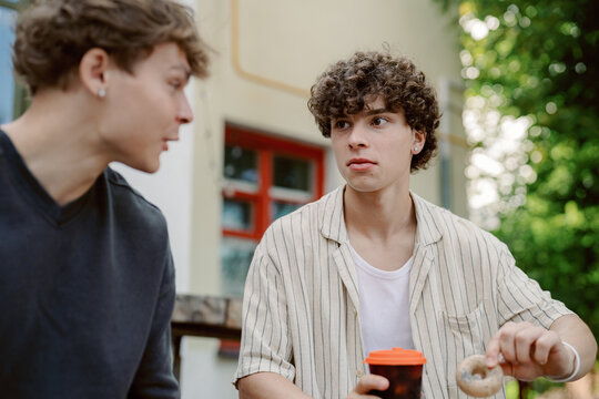 Two young men share a moment outdoors, engaging in lively conversation. One holds a cup of coffee while the other enjoys a donut, surrounded by greenery.