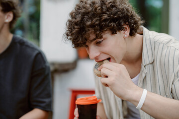 A young man with curly hair takes a bite of a pastry while holding a warm beverage. He is seated outdoors, dressed casually, and appears to be in a relaxed atmosphere with friends nearby.