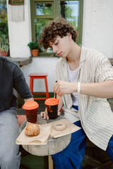 Two friends relax at an outdoor cafe, savoring beverages and treats. One young man focuses on his drink while seated at a wooden table decorated with pastries.
