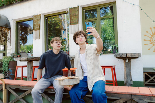 Two young men sit at a cafe, enjoying drinks and food. One takes a selfie with his phone while the other watches, surrounded by a vibrant outdoor setting with green plants.