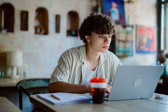 A young man with curly hair is focused on his laptop in a cozy cafe. He is surrounded by warm decor and has a drink beside him, creating a productive atmosphere.