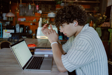 A young man with curly hair sits at a wooden table, deeply focused on his laptop screen. He wears earbuds and is surrounded by a cozy, colorful workspace filled with plants and decor.