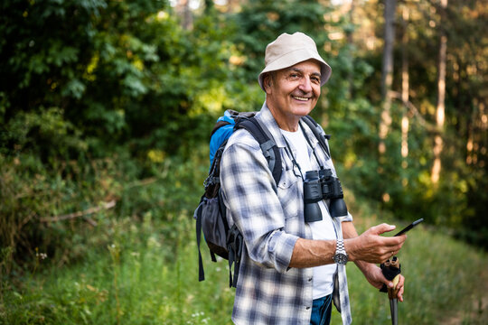Portrait of happy senior man with backpack and hiking poles using smartphone while enjoying hiking in nature.	