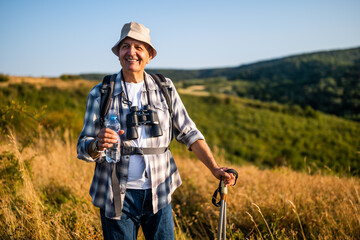 Portrait of smiling senior man drinking water while enjoying hiking with backpack and hiking poles in nature.	