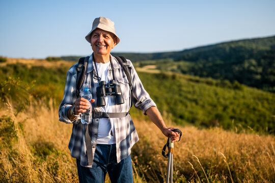 Portrait of smiling senior man drinking water while enjoying hiking with backpack and hiking poles in nature.	 - Powered by Adobe