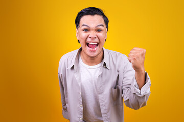 Excited Young Asian Male Celebrating Victory With Triumphant Fist Pump Isolated On Yellow Background