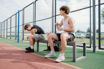 Two young male athletes are sitting on a bench at a sports facility, catching their breath after an...
