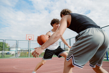 Two boys are engaged in a fast-paced basketball drill on an outdoor court. They showcase skills while dribbling and vying for position. Clear sky indicates a sunny day.
