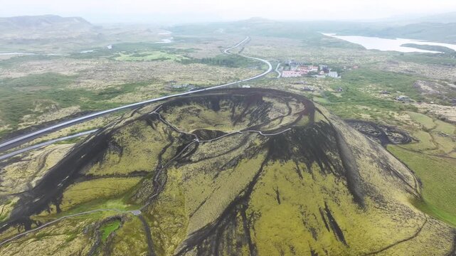 Green grabrok volcano crater on Iceland with traffic on road. Cloudy summer day. Aerial wide shot.