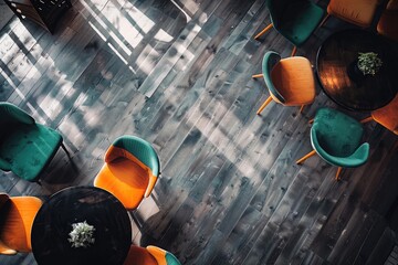High-angle view of a room with dark wood floors, small round tables, and orange and teal chairs arranged in a casual seating area. Sunlight streams in through a window, casting shadows on the floor