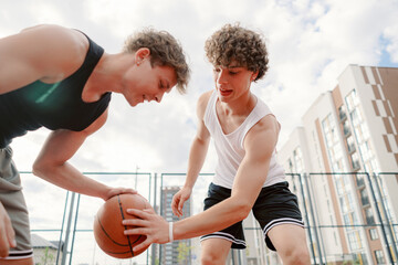 Two young male players engage in a competitive basketball game on an outdoor court, showcasing teamwork and skill. The sky is blue with scattered clouds, creating a vibrant atmosphere.