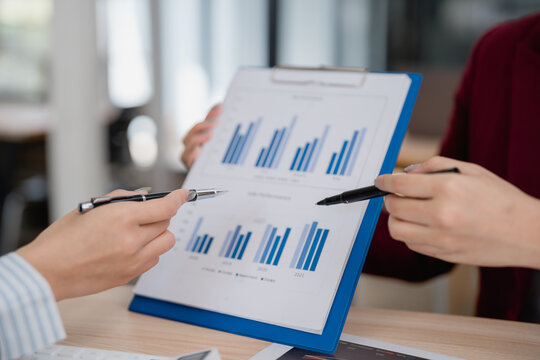 Two businesswomen are pointing at charts on a clipboard, analyzing financial data and discussing company performance during an office meeting - Powered by Adobe