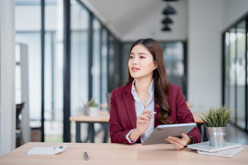 Young asian businesswoman holding tablet and stylus pen, thinking about new ideas for her business, sitting at desk in modern office, looking away