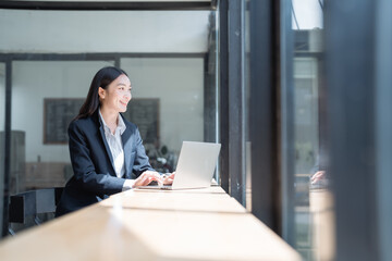 Young asian businesswoman sitting at desk in office, working on laptop computer and looking outside window, enjoying break time, smiling happy, natural light