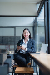 Young Asian businesswoman sitting at a sleek desk in a modern office, using her smartphone while working on her laptop, smiling confidently and engaging with the camera
