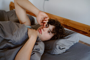 A young man with curly hair lies in bed, waking up and stretching. Natural light floods a cozy bedroom, creating a warm and inviting atmosphere during the morning hours.