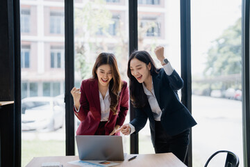 Two excited businesswomen raising their arms in celebration while gazing at a laptop in the office, reveling in the joy of achieving their professional goals and milestones