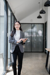 Young Asian businesswoman smiling while walking through a modern office corridor, using a digital tablet amidst bright glass walls, embodying professionalism and confidence
