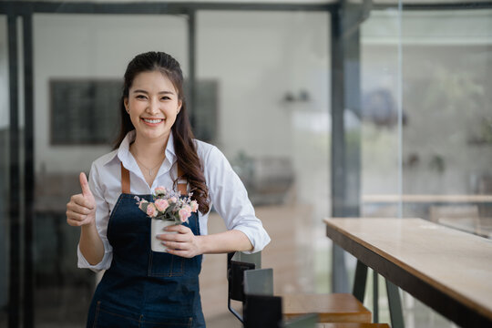 Young Asian waitress in an apron holding flowers and giving a thumbs up, with large windows in the background, embodying small business success and customer satisfaction