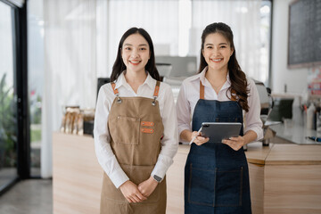 Two young, smiling Asian waitresses wearing aprons pose in their coffee shop, with one holding a tablet, radiating confidence and friendliness in a bright, welcoming atmosphere