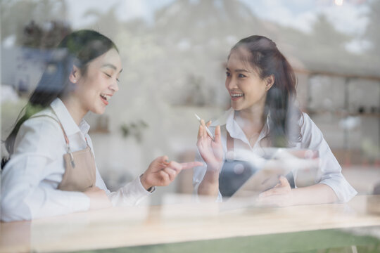 Two young, smiling waitresses discussing work schedules while using a tablet and digital pen, captured through a cafe window, highlighting teamwork and collaboration in a vibrant setting