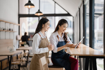 Two young, smiling waitresses wearing aprons are engaging with a tablet in a modern cafe, discussing their work tasks and planning the day ahead while enjoying the vibrant atmosphere