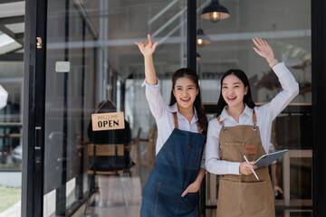 Two asian female cafe owners wearing aprons are smiling and waving to customers in front of their coffee shop with an open sign hanging on the door