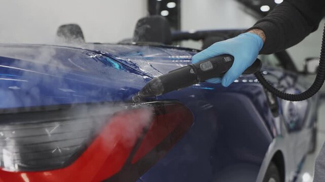 Professional automotive detailer applying protective film on blue car, using steam heat gun for precise wrapping and seamless paint protection