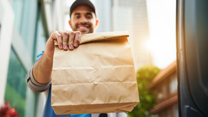 Smiling food delivery courier standing outdoors, holding out a brown paper bag with takeaway order towards the camera, with blurred city buildings and warm sunlight in the background