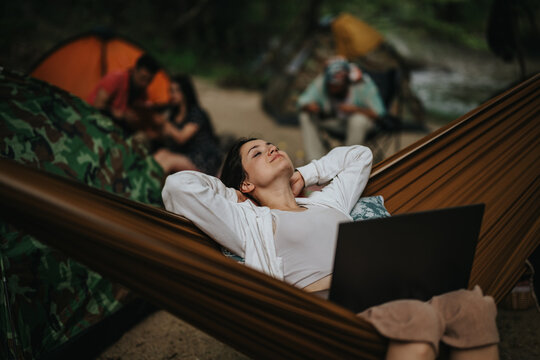 A carefree individual lounges in a hammock at a forest campsite while their friends enjoy the natural surroundings. Perfectly capturing relaxation, friendship, and the beauty of outdoor adventures.