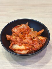 Authentic Korean Side Dish: Two Vertical Shots of Freshly Fermented Spicy Napa Cabbage Kimchi (Baechu Kimchi) in a Black Ceramic Bowl, Isolated on a Light Wooden Background.