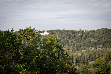 Obraz premium A scenic landscape showing a historic white mansion located on top of a forested hill, surrounded by lush green trees and natural woodland