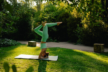 Young woman in sportswear standing on her head on fitness mat at public park, doing yoga. Healthy...