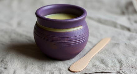 A handcrafted, purple ceramic pot, filled with a light-yellow liquid, sits on a gray linen tablecloth, accompanied by a wooden serving utensil.