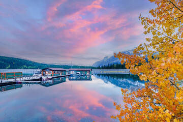 Lake Minnewanka, Canada: A peaceful lakeside vista where autumn's golden leaves meet towering Canadian Rockies, reflecting silently in the calm, clear glacial waters