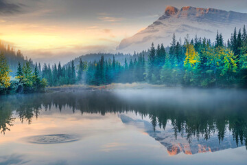 Beautiful Lake Minnewanka, Canada, showcasing tranquil water, evergreen trees, mountain peaks, and sunrise sky. Ideal for nature wallpaper, travel marketing, adventure tourism.