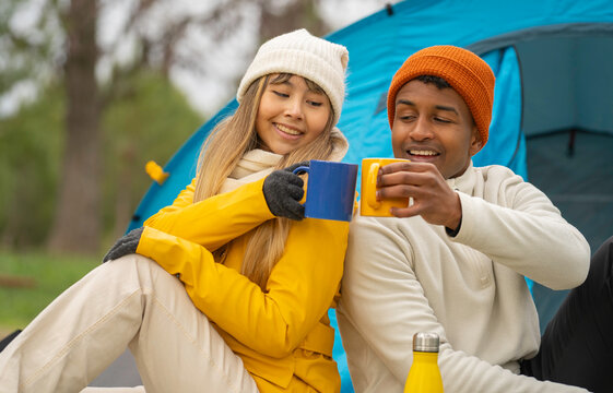 Interracial couple enjoying a camping trip, toasting with mugs in front of their tent