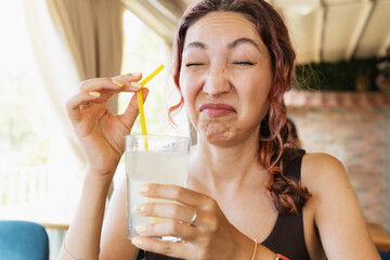 Young woman making a disgusted funny face while drinking a glass of sour lemonade with a straw in a restaurant