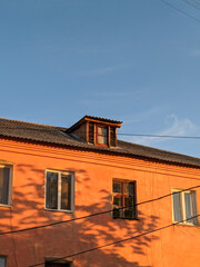 Old orange building with a wooden dormer window, illuminated by warm golden hour light. Tree shadows fall on the facade under a clear blue sky, creating a peaceful evening mood.