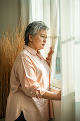 Senior Asian woman gazing through curtain in morning sun light with peaceful expression