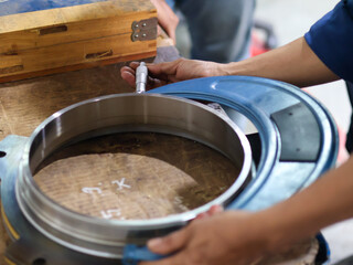 Close-up of a person's hands using a large Screw micrometer to measure a metal ring. The image captures the industrial process, focusing on precision, quality, and the tools involved