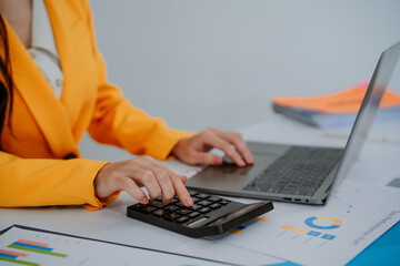 Asian businesswoman using laptop and analyzing financial charts, holding a calculator and working with graphs in modern office, smiling and focused on her tasks