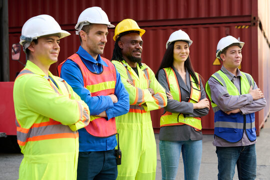 A diverse group of shipping workers with arms crossed, in front of container equipment. They have helmets and safety vests